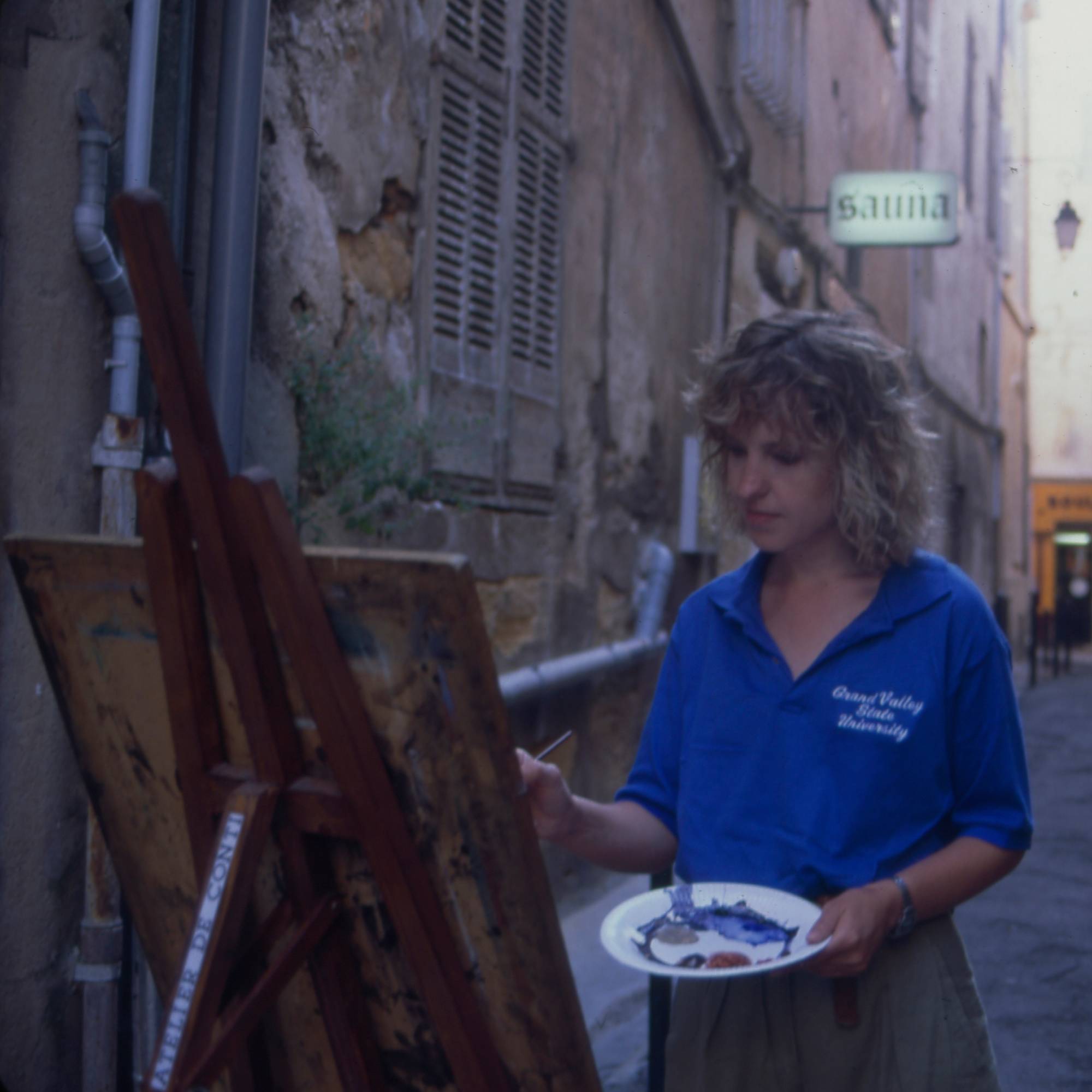 young woman in blue GVSU polo shirt painting in cobblestone street in France